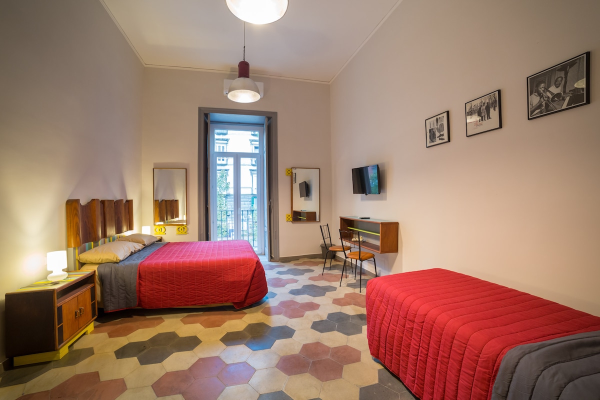 A spacious room features a double bed and a single bed, both dressed in red bedding. High ceilings and large windows allow natural light, while framed black-and-white photographs adorn the walls. A small desk and chair are positioned against one wall, enhancing the functional layout.