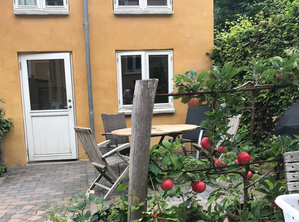 A bright yellow exterior of the guesthouse is showcased, featuring a wooden door and two windows. In the foreground, a wooden table surrounded by four chairs offers a space for outdoor gatherings. A garden with apple trees and green foliage enhances the setting.