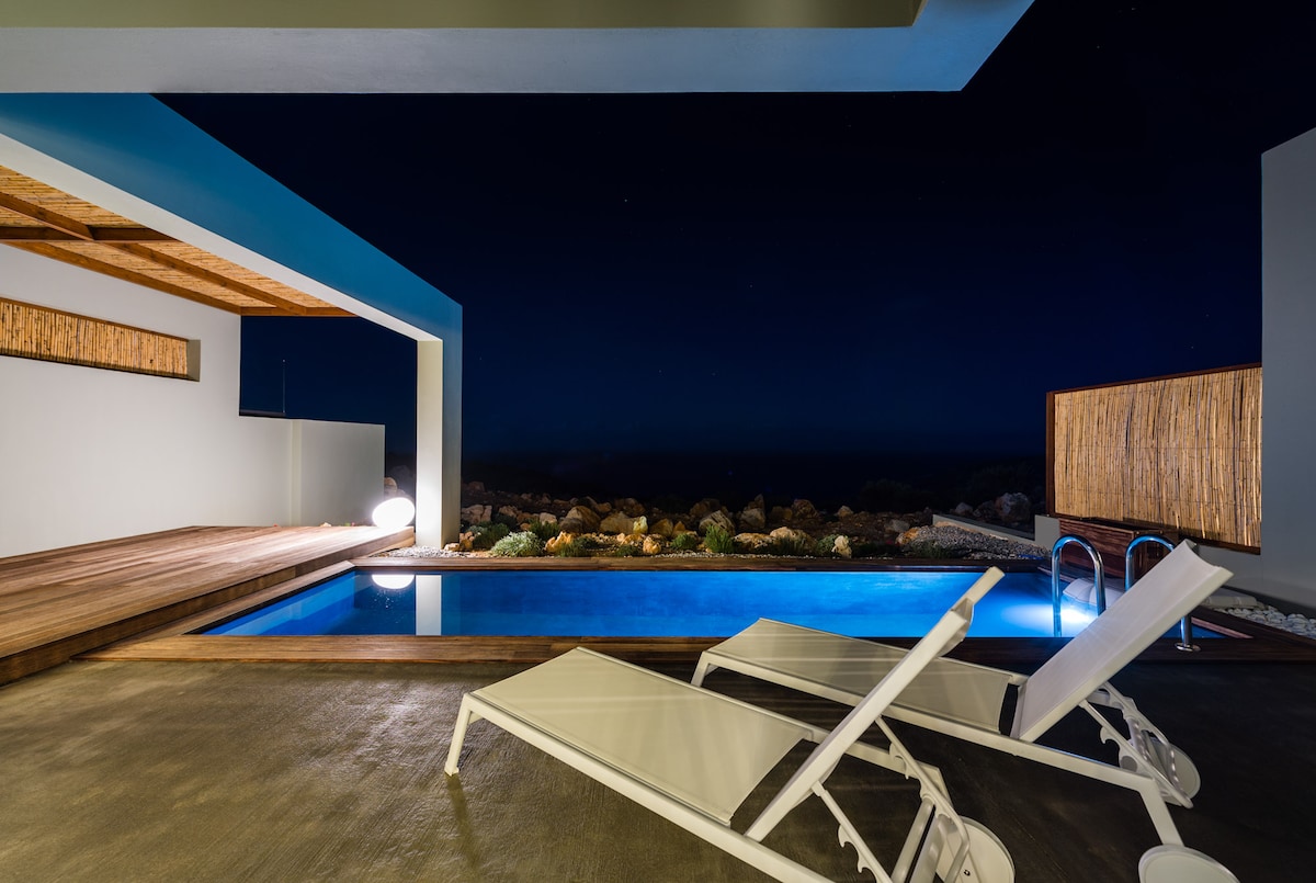 A modern outdoor space is featured, showcasing a private pool with vibrant blue water under the night sky. Two white lounge chairs are positioned by the poolside, with natural landscaping visible in the background, enhancing the tranquil atmosphere of the setting.
