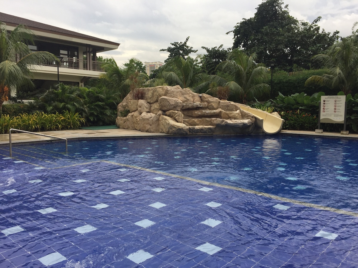 A spacious outdoor pool features a textured rock formation with a slide, surrounded by lush greenery and well-maintained landscaping. The water surface reflects a vibrant blue hue, while tiles with varied patterns are visible along the pool's edge.