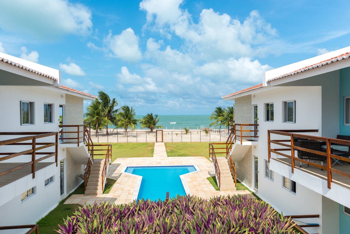 The image captures a serene view of two buildings flanking a central pool area. Lush greenery lines the walkway leading to the beach, while the ocean glistens under a sunlit sky, enhancing the overall coastal ambiance.