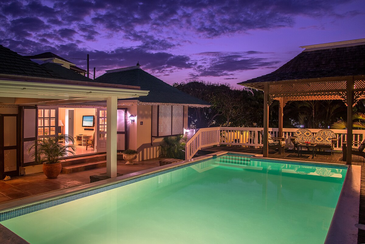 The private pool glows in soft turquoise, surrounded by a tasteful deck space and gazebo. The setting sun casts a warm purple hue across the sky, while cozy seating options offer a peaceful view of the lush landscape.