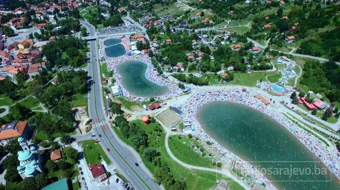 An aerial view captures the vibrant scene of Panonska Jezera, showcasing two large bodies of water surrounded by sandy beaches. Numerous sunbathers are visible along the shore, while greenery and residential areas form the backdrop. The nearby road features a steady flow of vehicles.