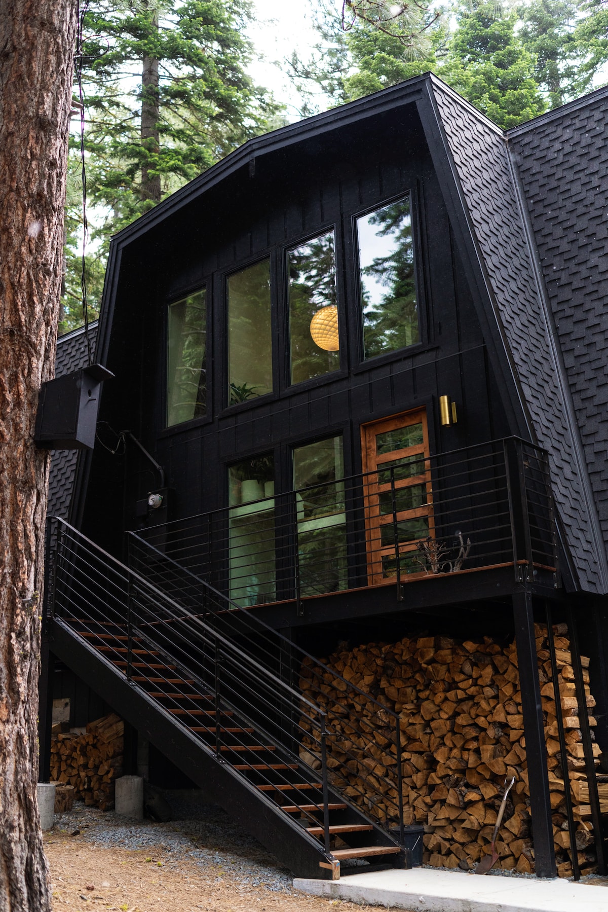 The exterior of the Black House Hideaway is characterized by a striking black façade and angled rooflines. Large windows provide ample natural light, while a staircase leads to the entrance. A stack of firewood is neatly arranged beneath, enhancing the rustic aesthetic.