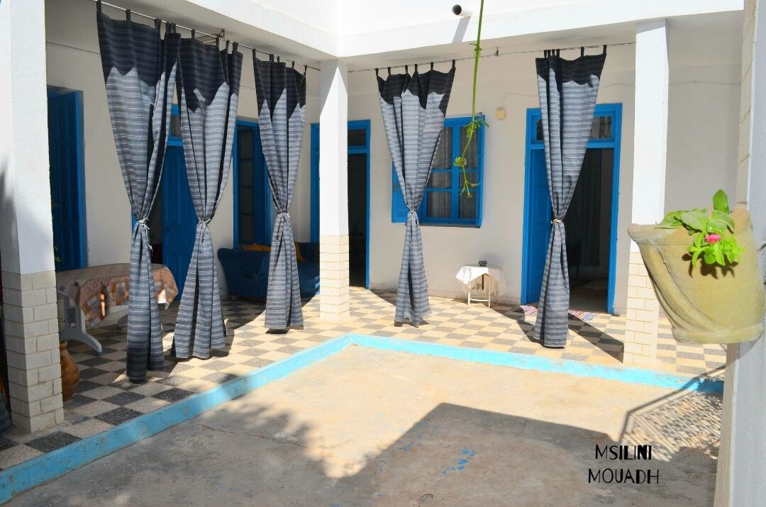 An open courtyard is framed by tall, dark curtains, providing a sense of privacy. Vibrant blue doors are visible in the background, complementing the tiled floor pattern. A small plant is situated in the corner, adding a touch of greenery to the space.