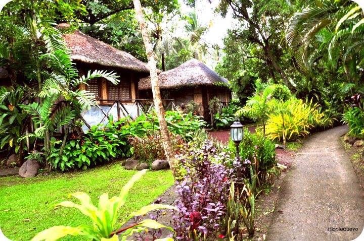 A serene pathway meanders through lush greenery, leading to charming cottages with thatched roofs. Vibrant tropical plants and flowers adorn the landscape, creating a tranquil atmosphere. Soft lighting from a lantern enhances the inviting setting amidst the natural surroundings.