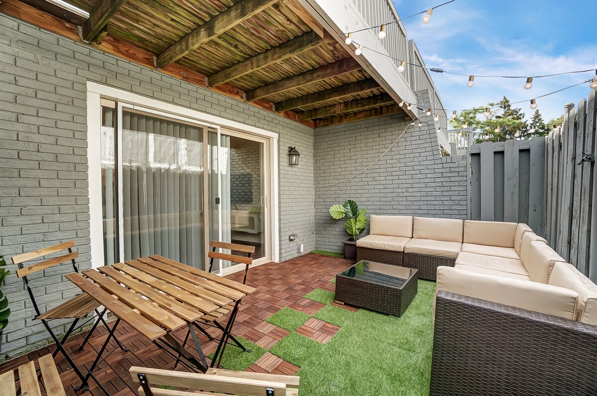 A private patio features a comfortable seating area with a sectional sofa, complemented by a coffee table. A dining table with folding chairs is set against a backdrop of greenery, while string lights enhance the outdoor ambiance. Sliding glass doors provide access to the apartment.