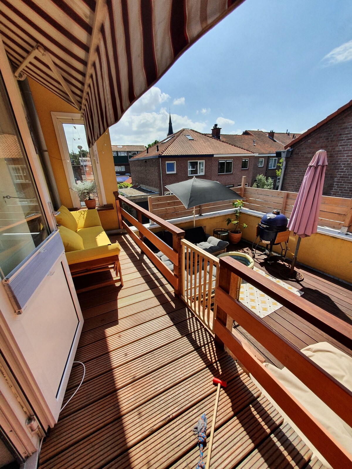 A spacious terrace features wooden decking, with a sun umbrella and seating area inviting relaxation. Potted plants and a barbecue area are visible, while surrounding rooftops and a church steeple provide a backdrop, creating an open and airy atmosphere.