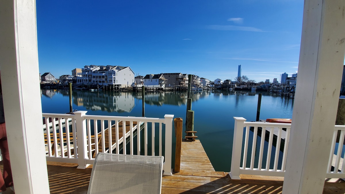 A serene waterway reflects clear blue skies and surrounding buildings. Wooden docks extend into the water, bordered by white railings. Several boats are visible nearby, enhancing the tranquil setting.