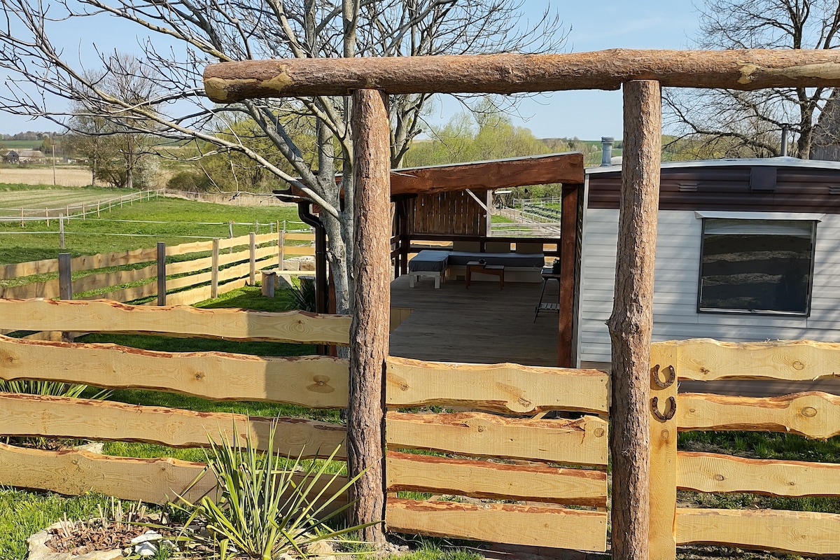 A rustic wooden fence surrounds a spacious backyard with a canopy-covered seating area. The entrance gate, crafted from logs, opens to a serene landscape of grassy fields and distant trees, enhancing the peaceful atmosphere of the rural setting.