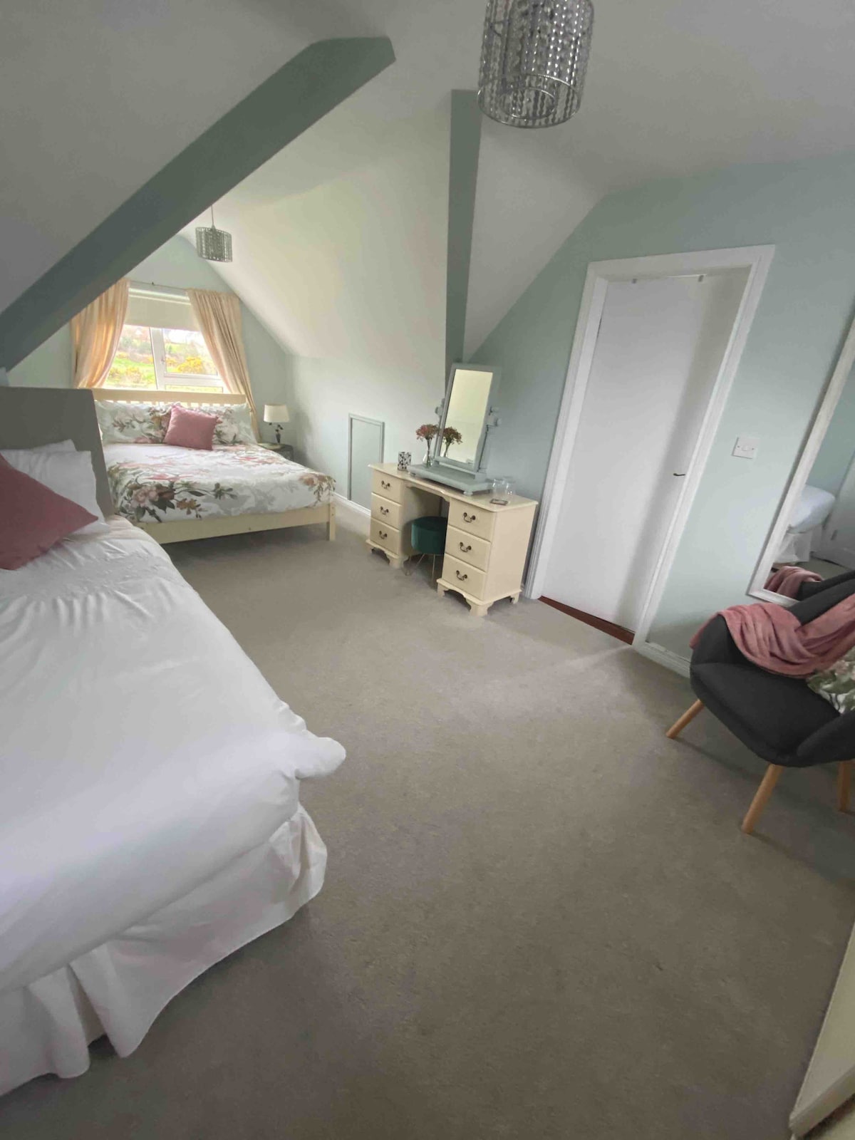 A cozy bedroom is presented with two beds, one spacious and the other a smaller twin. Soft natural light enters through a window adorned with sheer curtains. A dresser and a mirror are positioned against the wall, complemented by a small chair and a plaid throw.