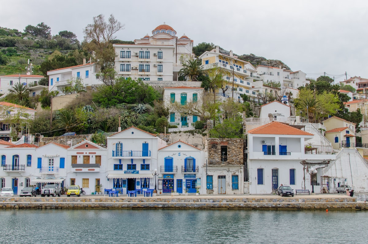 A view of the traditional village of Evdilos is shown, highlighting the colorful buildings with blue accents along the waterfront. The gently sloping hillside is lined with greenery, while pedestrians can be seen along the scenic path. The cloudy sky adds a serene atmosphere.