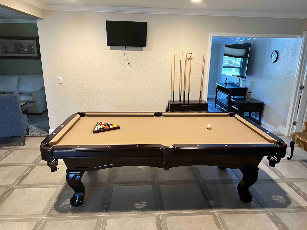 A billiard table is centered in a well-lit game room, equipped with cues positioned in a rack nearby. A flat-screen TV is mounted on the wall, with light-colored walls contributing to the space's brightness. The room is connected to a view of the living area through an open doorway.