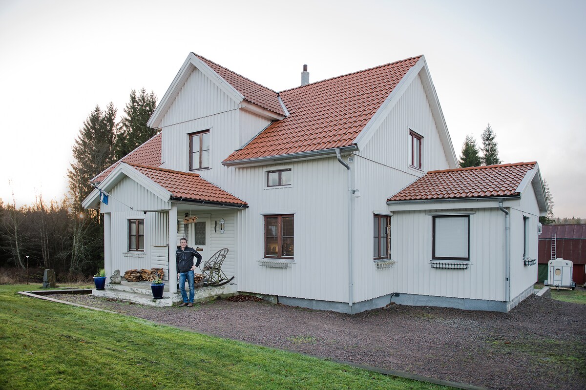 A two-story house constructed with light-colored wood features a red-tiled roof. Multiple windows are present, allowing natural light to enter. A person stands near the entrance, with a lawn surrounding the property, providing open space and a tranquil atmosphere.