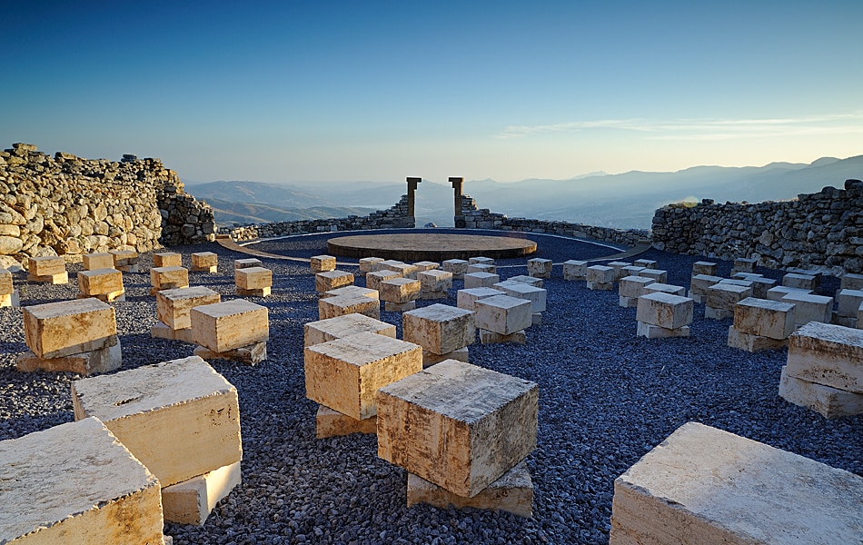 An open-air archaeological site features a circular arrangement of stone blocks on a gravel surface. In the background, ancient walls are partially visible, framing a panoramic view of rolling hills under a clear sky at dawn.
