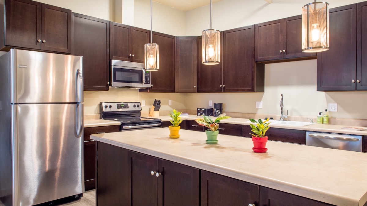 A modern kitchen features dark cabinetry and stainless steel appliances. A spacious island is highlighted by three small potted plants, which add a touch of greenery. Pendant light fixtures hang above, providing warm illumination over the countertop and creating a functional cooking space.