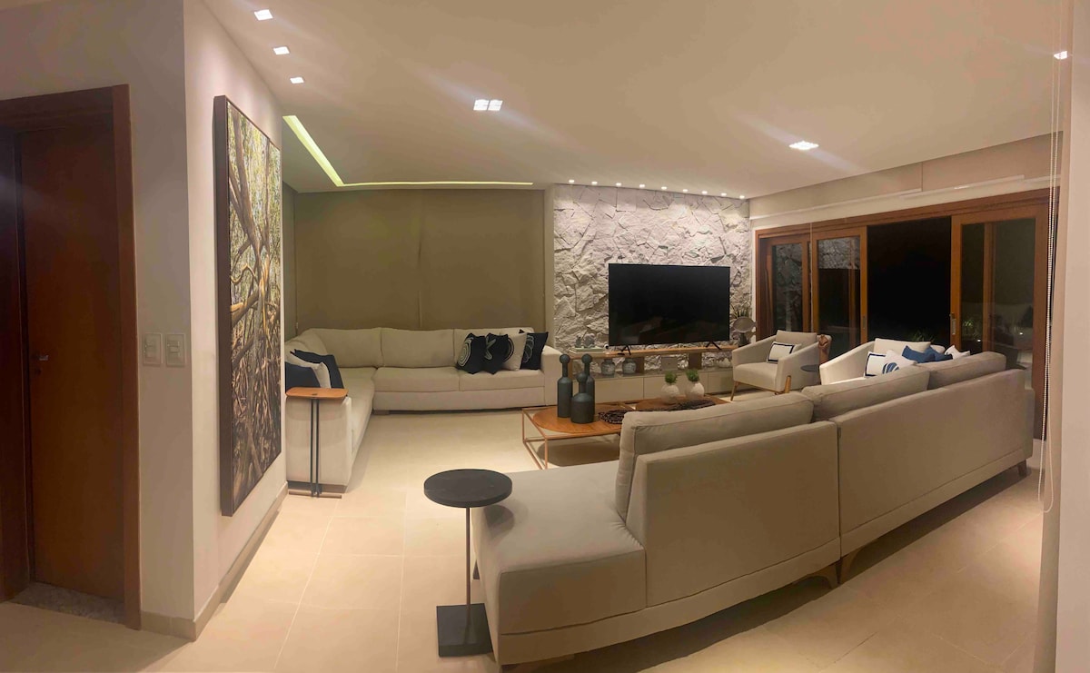 A spacious living area is highlighted by a large sectional sofa adorned with decorative pillows. A contemporary coffee table sits in front, with a low side table positioned to one side. A wall-mounted television is framed by decorative stonework, and large windows offer a view of the outdoors.