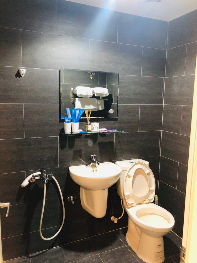 A compact bathroom displays a modern design with dark tiled walls. A white toilet is positioned adjacent to a small sink, which is accompanied by a shelf holding toiletries. A handheld showerhead is mounted on the wall for convenience.