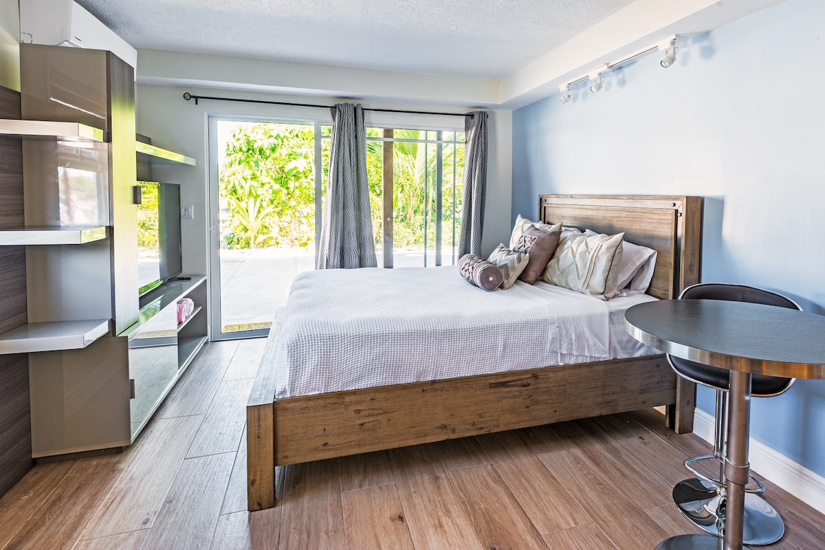 A spacious studio unit features a wooden queen bed with a light blanket and decorative pillows. Natural light enters through a set of glass doors, leading to a private patio. A small round table with bar stools complements the minimalist decor.
