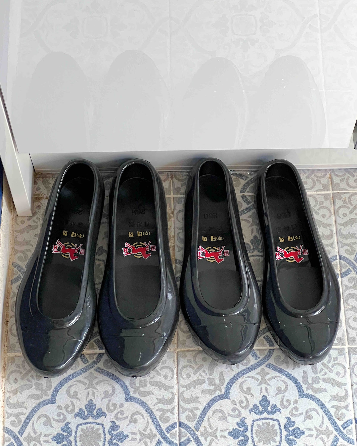 A pair of sleek black flats is arranged side by side on a patterned tiled floor. The shoes feature a glossy finish and are designed for comfort. Light reflects off their surfaces, highlighting their smooth contours.