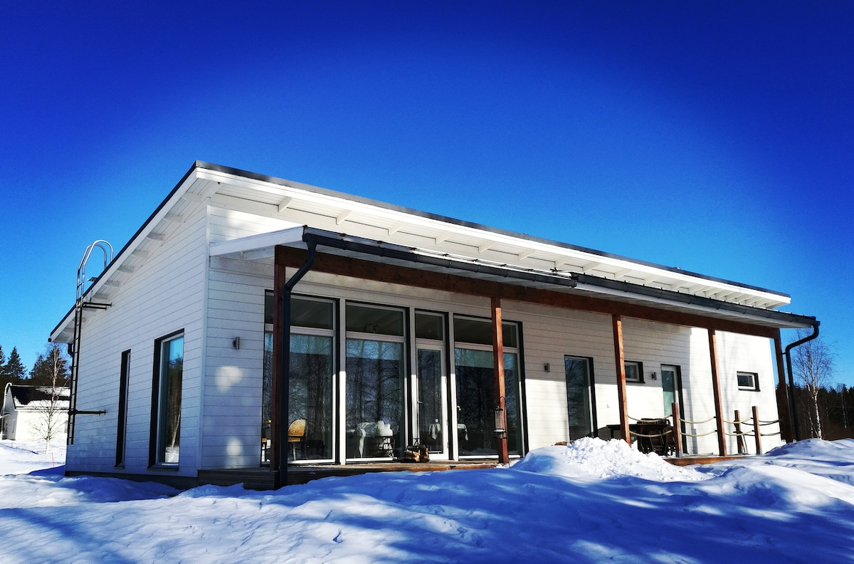 A modern single-story house is depicted with large glass doors and windows, allowing natural light to enter. The snow-covered ground surrounds the property, while a clear blue sky creates a serene backdrop. The structure features a covered terrace for outdoor seating.