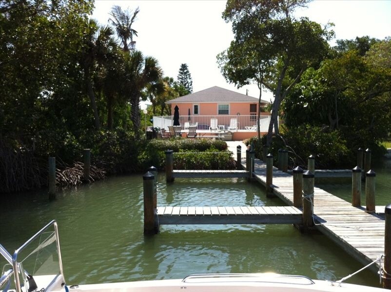 A serene view of the property from the water showcases a dock leading to a pastel-colored building surrounded by lush vegetation. Outdoor seating is visible on the deck, providing a peaceful place to relax under the sun.