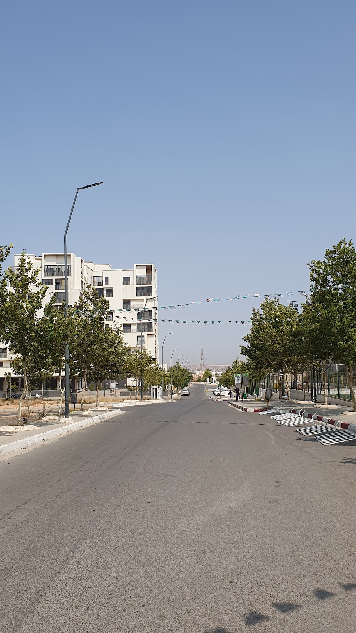 A wide street lined with trees extends toward a clear blue sky. Modern buildings are visible on either side, with decorative flags strung across the road. The pavement appears well-maintained, and the scene conveys a peaceful atmosphere in a suburban setting.