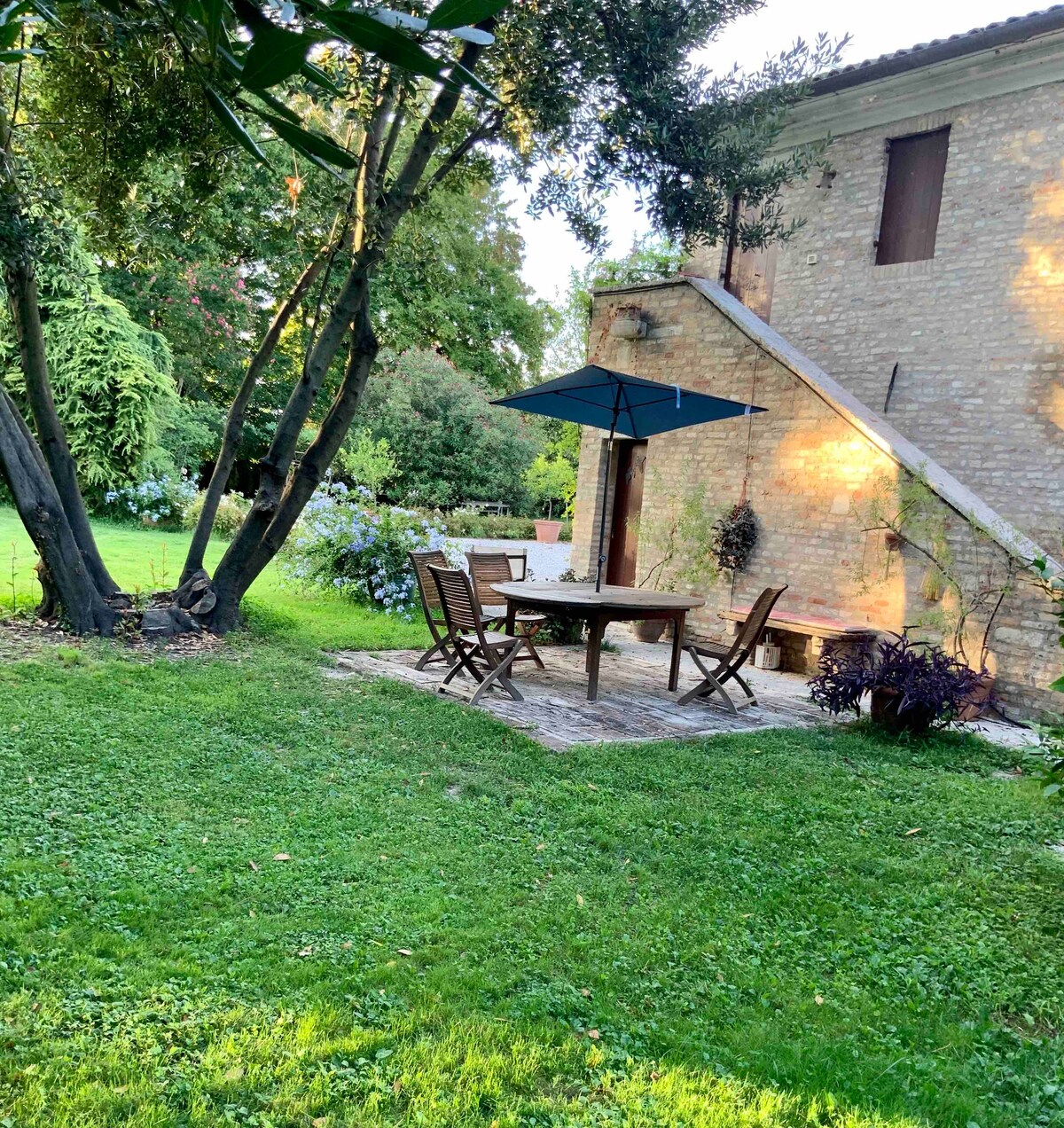 A shaded patio area features a wooden table surrounded by chairs, with an umbrella providing cover. Lush green grass and various plants create a serene outdoor environment, complemented by the stone wall of the building in the background.