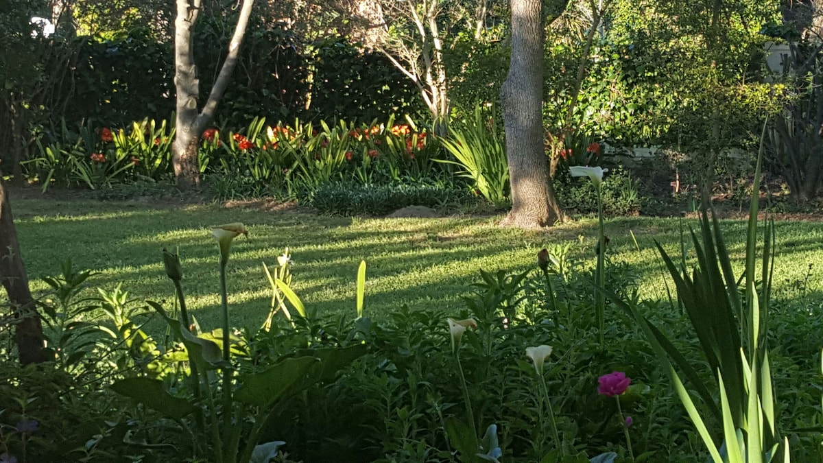 A landscaped garden area is showcased, featuring various plants and vibrant flowers. The lush green grass leads to flower beds with colorful blooms, while trees provide shade. Sunlight filters through the foliage, creating a serene outdoor setting ideal for relaxation.