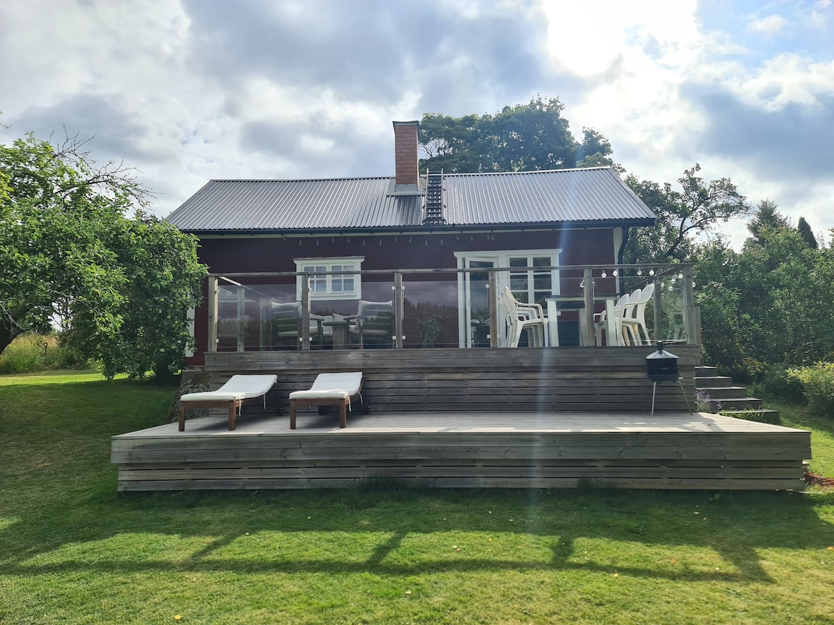A traditional wooden cabin is shown from the garden, featuring a spacious deck. Sun loungers and a small grill are positioned on the deck, while a set of white chairs and a table are visible in front of the cabin. Lush green grass surrounds the area.