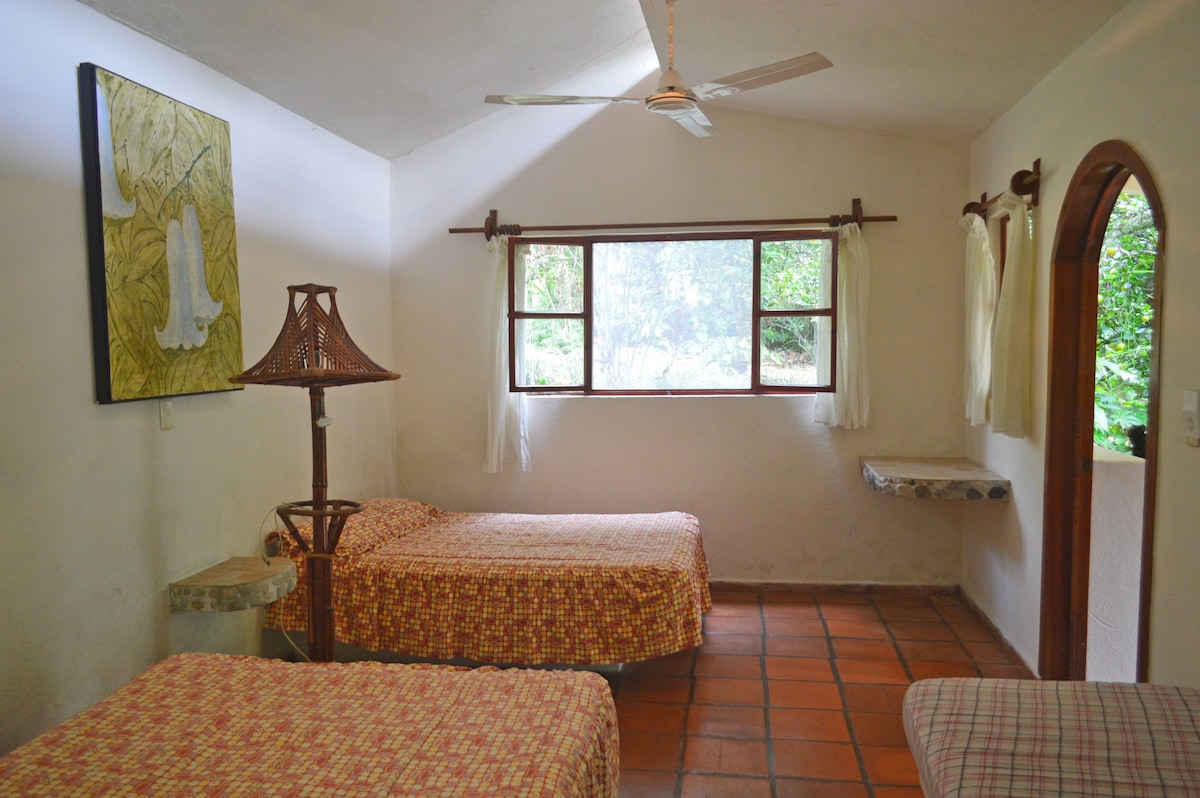A bright room features two spacious beds adorned with patterned bedspreads. Large windows allow natural light to illuminate the space, with views of the surrounding greenery. A decorative lamp and a small stone shelf add character to the room.