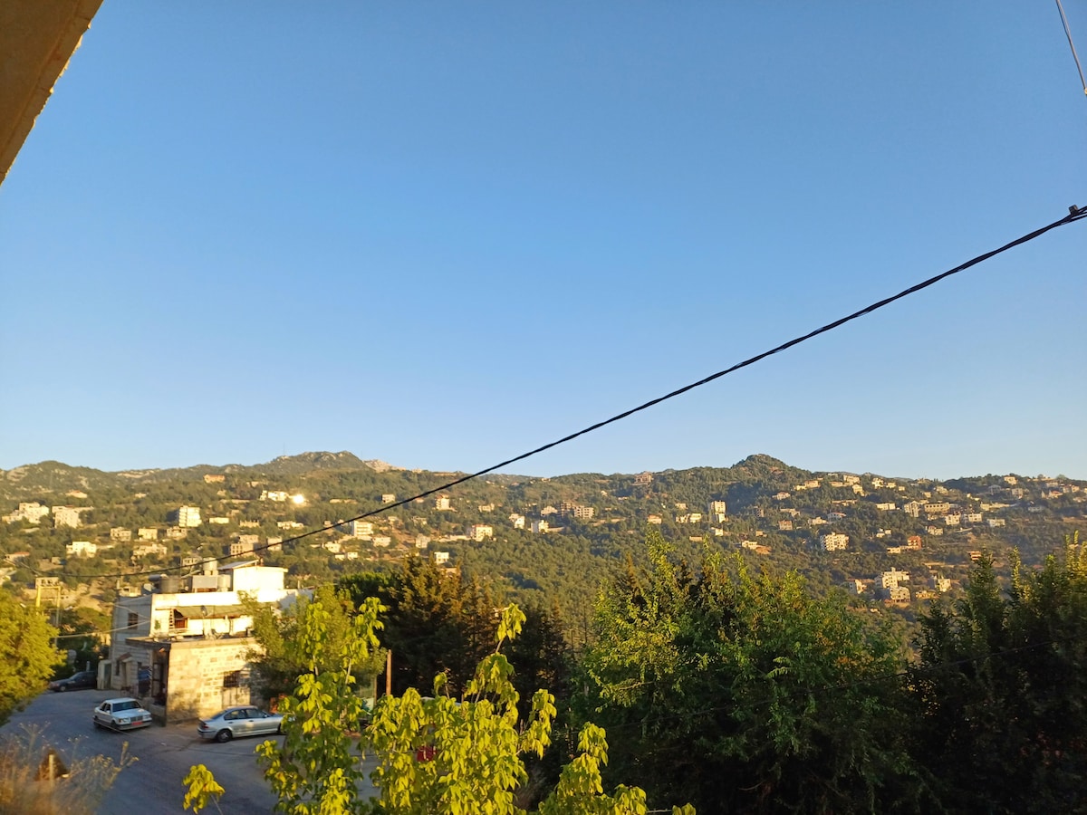 A wide view of rolling green hills under a clear blue sky is presented. The landscape is dotted with small white buildings, surrounded by trees. The serene atmosphere showcases the natural beauty of the village setting, allowing for a sense of calm and tranquility.