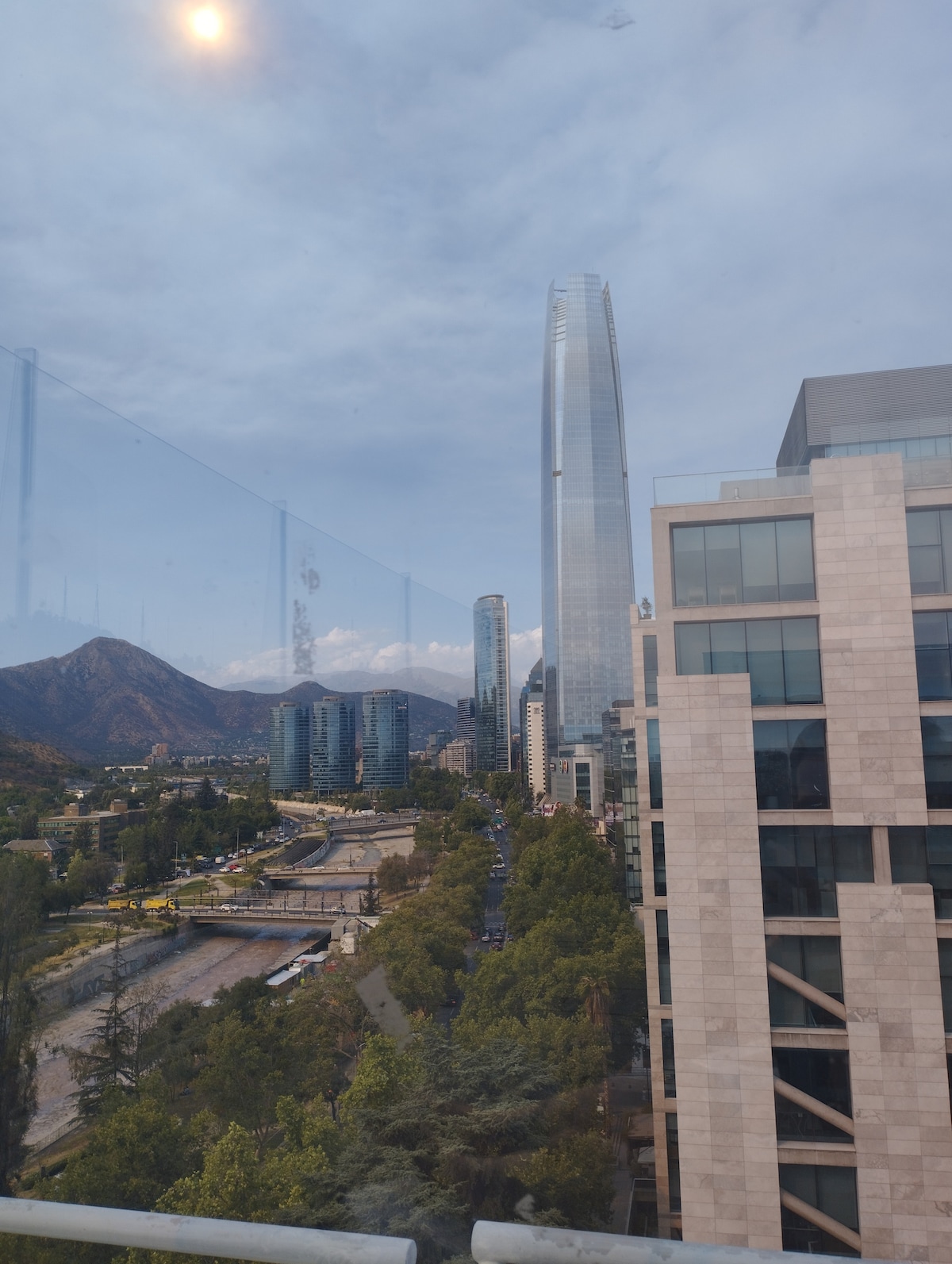 A panoramic view captures the skyline of Santiago, featuring the iconic Gran Torre Santiago towering amidst a backdrop of mountains. The image showcases a blend of modern architecture with urban greenery, creating a vibrant cityscape, framed by a glass barrier.