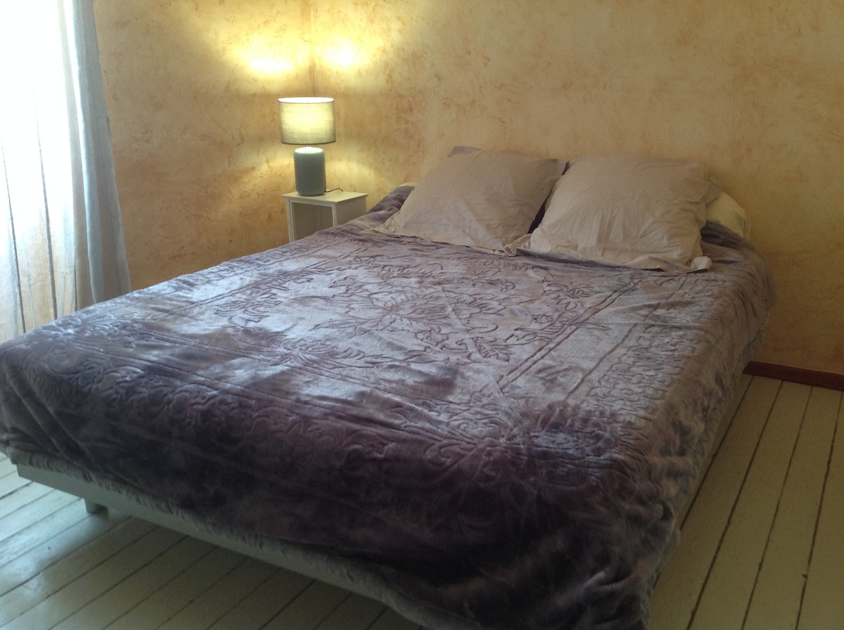A spacious bed is dressed in a soft, textured blanket, complemented by light-colored pillows. A bedside lamp casts a warm glow, while sheer curtains allow natural light to softly illuminate the room's textured walls.