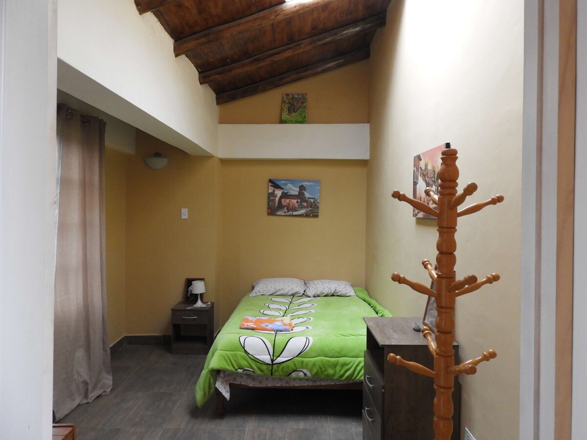 A cozy room with a green bedding set is highlighted by a high wooden ceiling. A bedside lamp and a small dresser are visible, along with a wooden coat rack standing in the corner. Natural light enters through the window, enhancing the room's brightness.