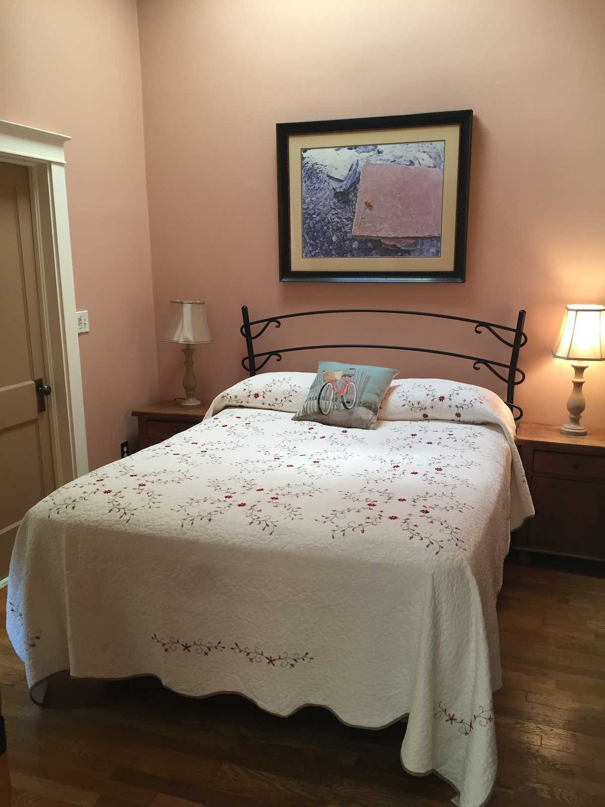 A cozy bedroom features a metal bed frame with a quilted coverlet adorned with floral patterns. Nightstands on either side hold soft-lit lamps. A framed artwork hangs above the bed, adding a touch of color to the pale pink walls, enhancing the room's serene ambiance.
