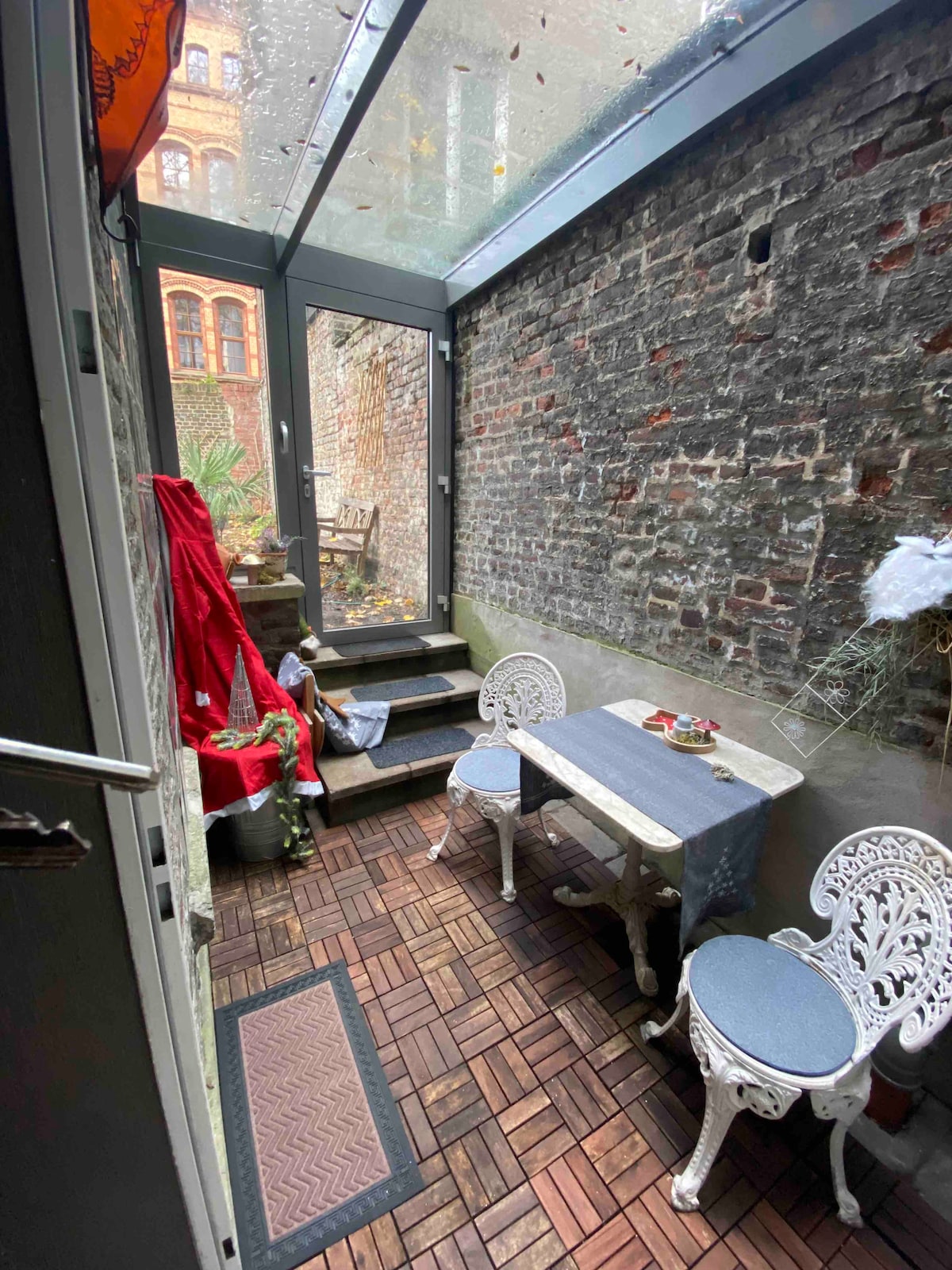 A small winter garden features a wooden floor and brick walls, with a set of two white chairs and a table. An intimate atmosphere is created by natural light filtering in through the glass roof. A few decorative items add a personal touch to the space.