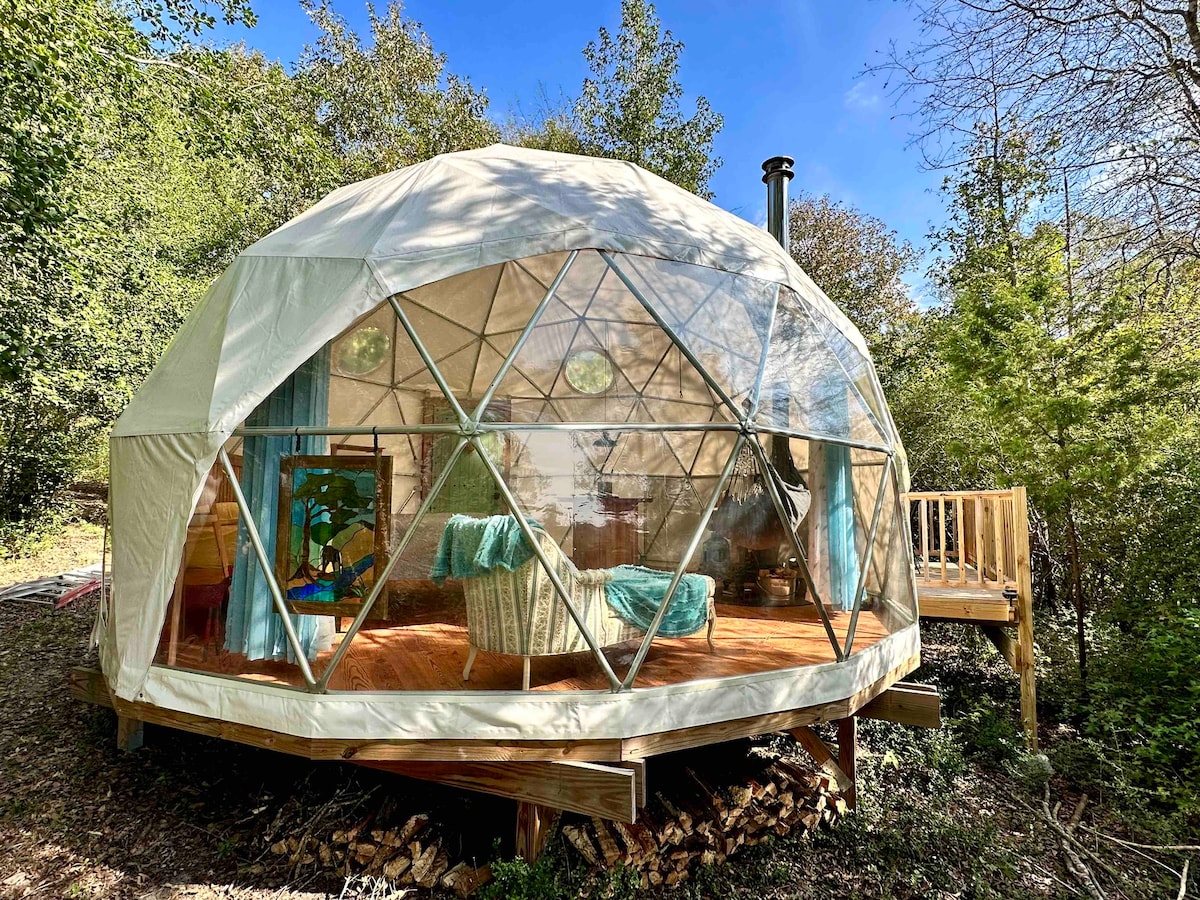 A unique Pacific dome structure is surrounded by lush greenery, showcasing a spacious deck. The dome features numerous geometric windows that allow natural light to illuminate the interior. A cozy chair is placed inside, complemented by soft furnishings, offering a comfortable retreat within the natural setting.