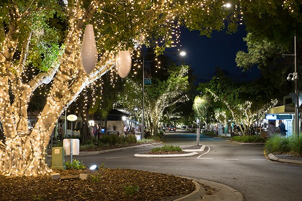 A charming street scene is captured at night, with trees adorned in warm, twinkling lights. The softly lit pathway guides the view, creating a serene atmosphere, while shops are subtly visible in the background, enhancing the inviting nature of the surroundings.