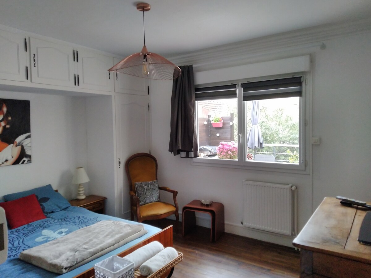A bright sleeping area includes a spacious bed adorned with a light blanket. A vintage armchair and small wooden side table complement the decor. Natural light enters through a large window, framed by light curtains and revealing a view of greenery and flowers outside.