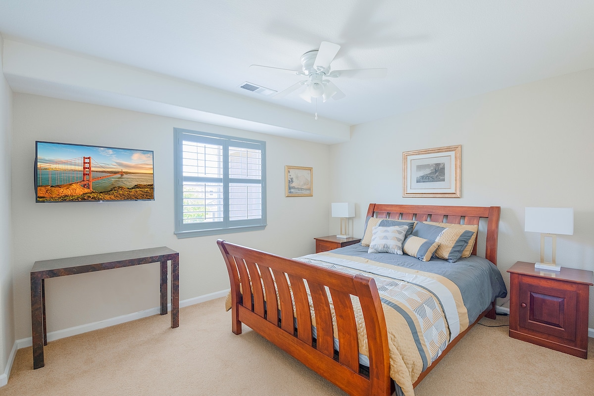 A bright bedroom features a wooden bed with a quilted coverlet. Nightstands with lamps flank each side. A flat-screen TV is mounted on the wall, and a window allows natural light to enter. Light neutral tones create a serene atmosphere.