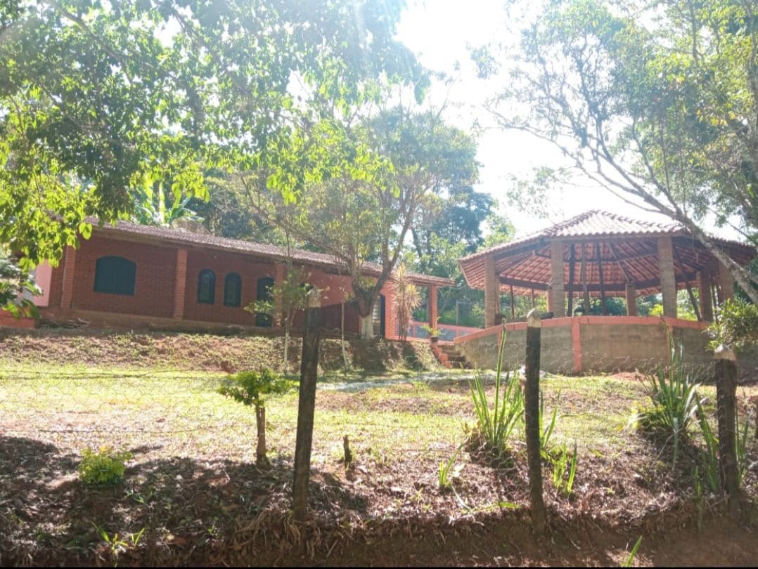 A serene outdoor view showcases a rustic brick building alongside a spacious covered area with a thatched roof. The setting is surrounded by lush greenery, with a grassy yard that invites relaxation. Sunlight filters through the trees, creating a calm ambiance.