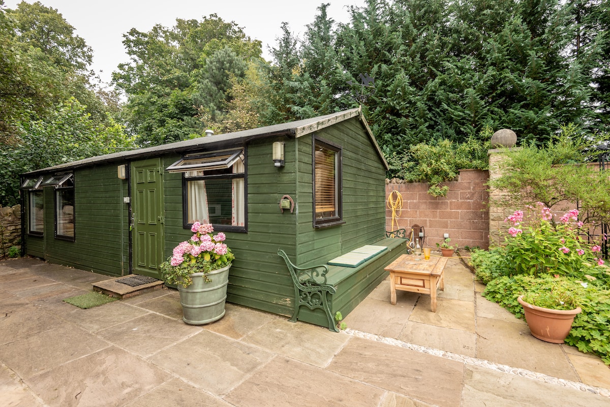 A detached green cottage is framed by lush greenery and colorful flowers. A small patio is visible, featuring a wooden table and chairs, alongside potted plants. The entrance is marked by a double door, with large windows allowing natural light to fill the space.