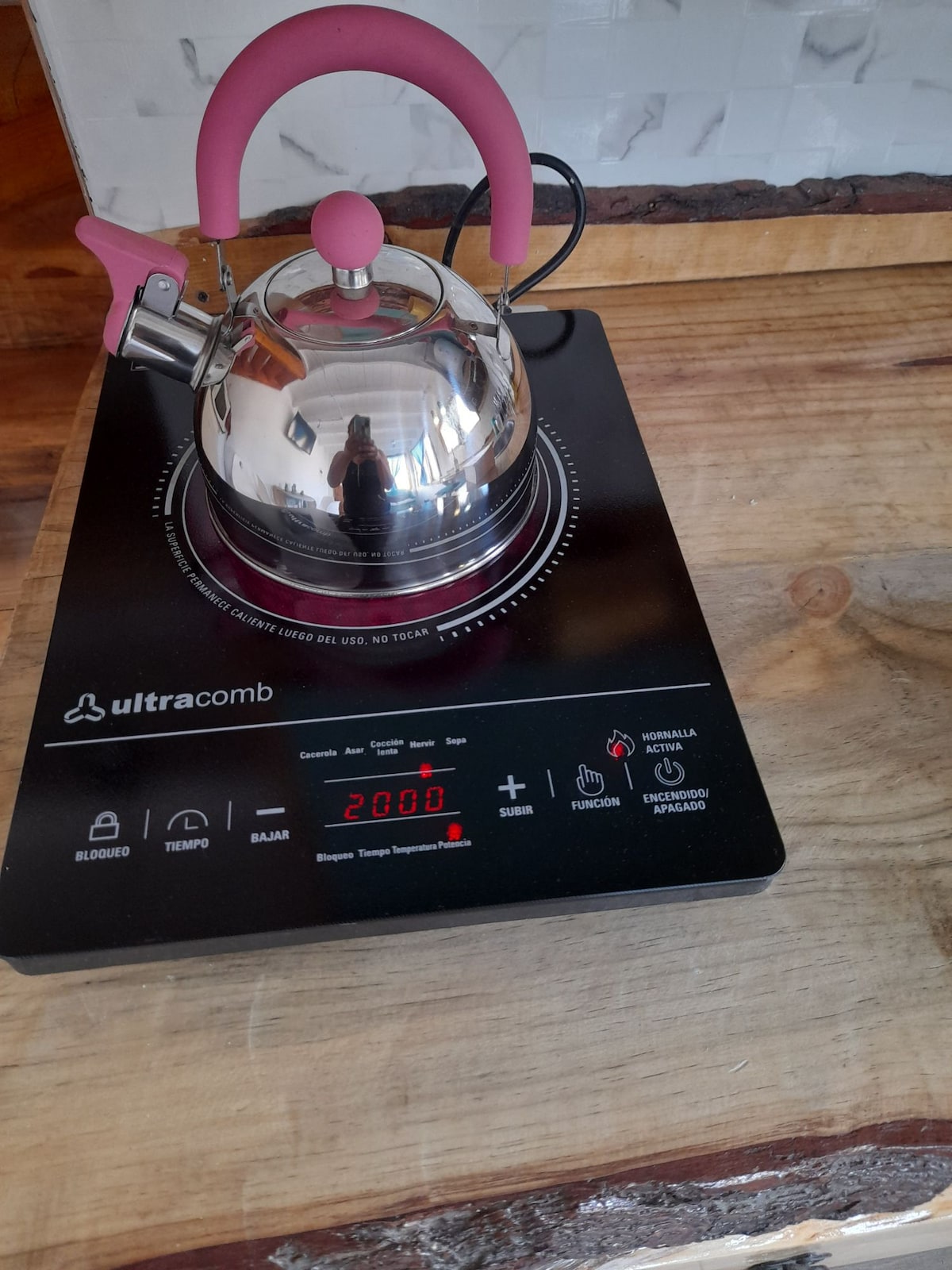 A sleek electric kettle with a pink handle is placed on a black induction stove. The kettle's shiny surface reflects its surroundings. The stove features a digital display with red numbers indicating the temperature settings, set on a natural wooden countertop.