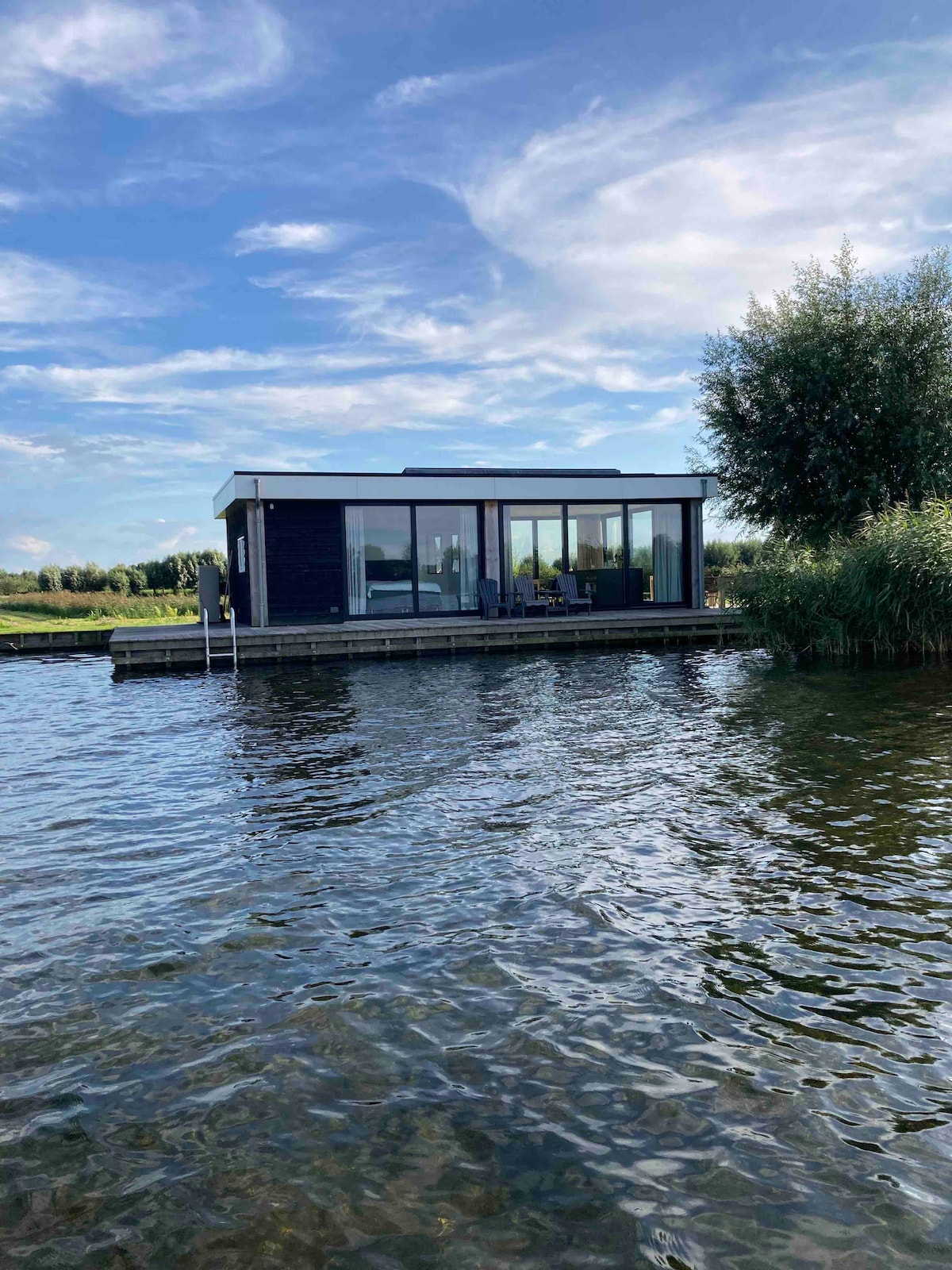 A modern house is situated on the water's edge, featuring large glass windows that reflect the sky. Surrounding greenery enhances the natural setting, while a small deck is visible alongside the structure. The serene water surface ripples gently in the foreground.