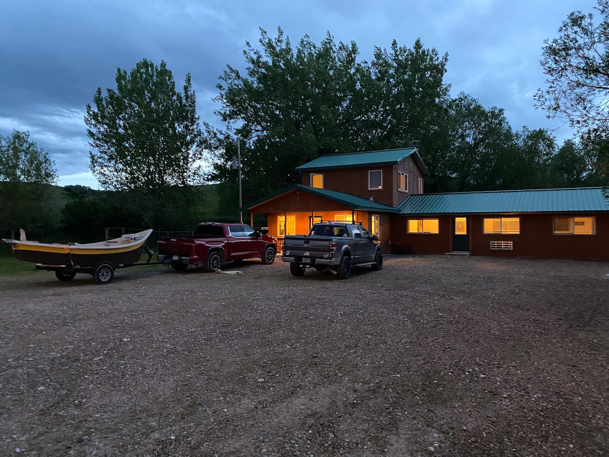 The exterior view of the Main Lodge is shown at dusk, featuring a brown wooden structure with a green metal roof. Several vehicles are parked in the gravel driveway, and multiple windows emit a warm glow, surrounded by trees in the background.