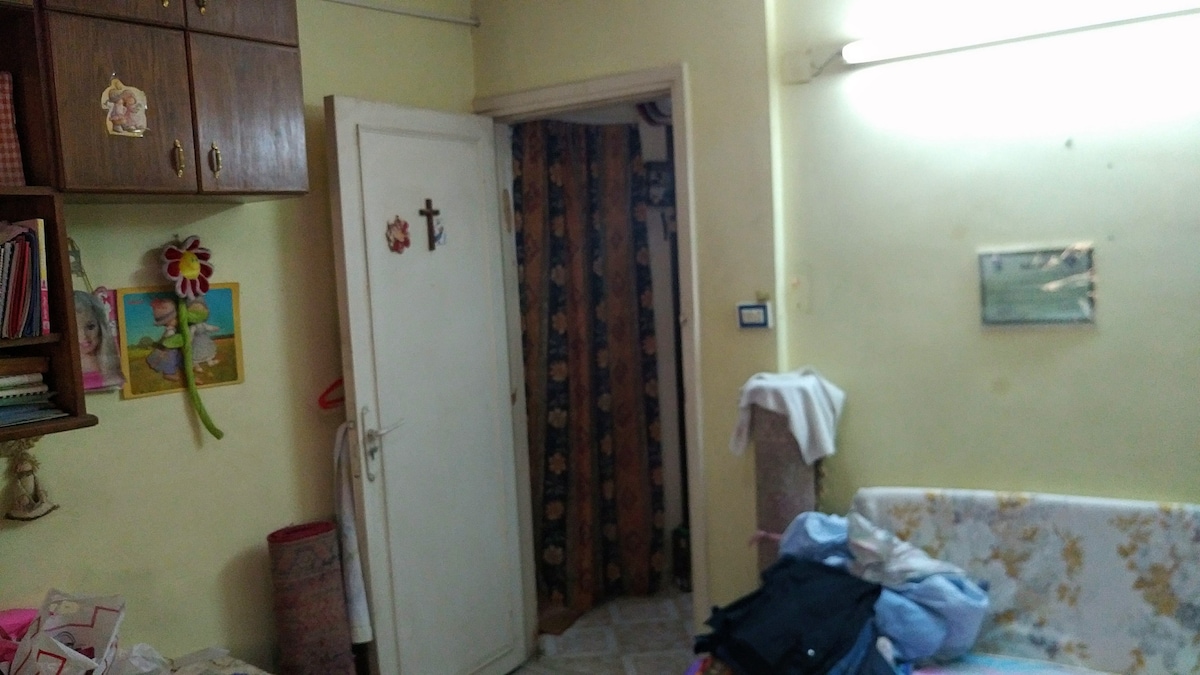 A cozy room features a light yellow wall with wooden cabinets displaying personal items. A closed white door with decorative accents leads to another space. A wall-mounted light illuminates the area, while a floral-patterned couch is adorned with assorted fabrics.