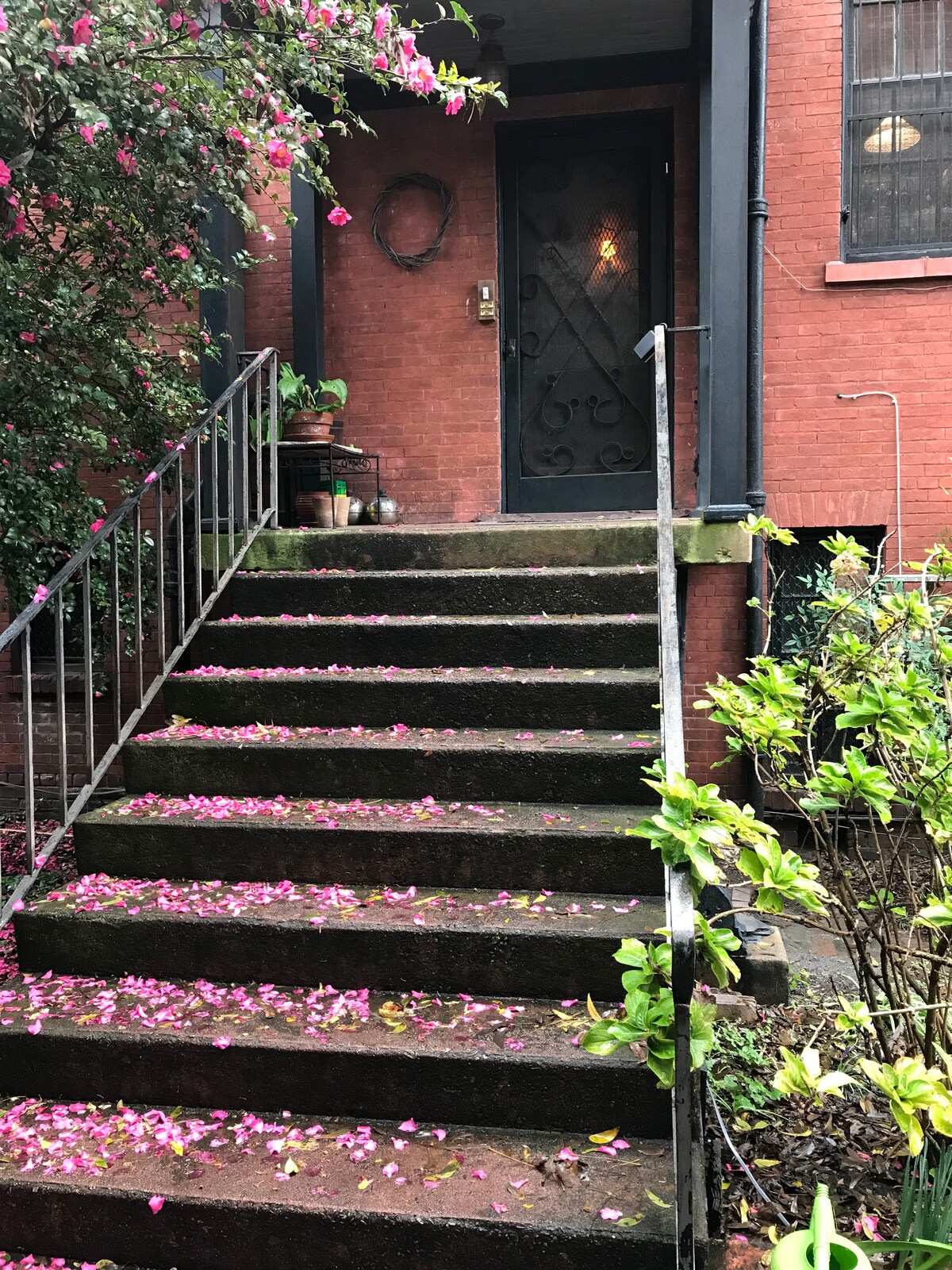 A charming entrance to the apartment suite features a set of stone steps adorned with scattered pink flower petals. The textured walls of the brick building create a warm backdrop, while a solid black door with a decorative design is illuminated by a soft light.
