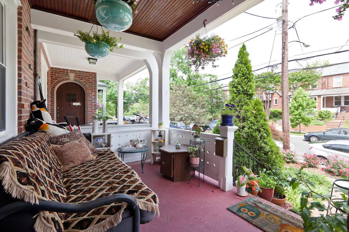 A covered porch features a cozy seating area with patterned cushions and decorative pillows. Hanging plants and potted flowers add color to the space, while a small desk stands against the railing. The porch provides views of the surrounding neighborhood and greenery.