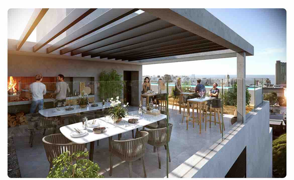 An outdoor terrace is showcased, featuring a spacious dining area with multiple tables and chairs. A built-in grill is visible, along with lush greenery surrounding the space. The skyline of Montevideo can be seen in the background, with sunlight illuminating the area.